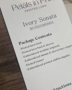 Product label for Roheenaz Ivory Sonata from the Petals in Prints printed lawn collection, showing package contents and a folded plaid fabric swatch on a wooden surface